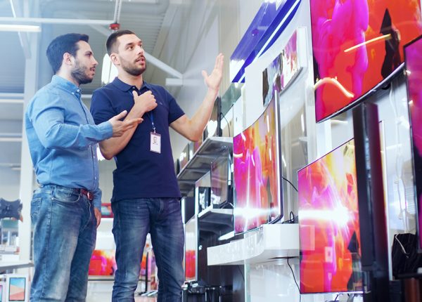In The Electronics Store Professional Consultant Shows Latest 4K Uhd Tv'S To A Young Man, They Talk About Specifications And What Model Is Best For Young Man'S Home. Store Is Bright, Modern And Has All The Latest Models.