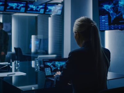 Fraud Special Agent Works on a Laptop in the Background Special Agent in Charge Talks To a Military Man in the Monitoring Room. In the Background Busy System Control Center with Monitors Showing Data Flow.